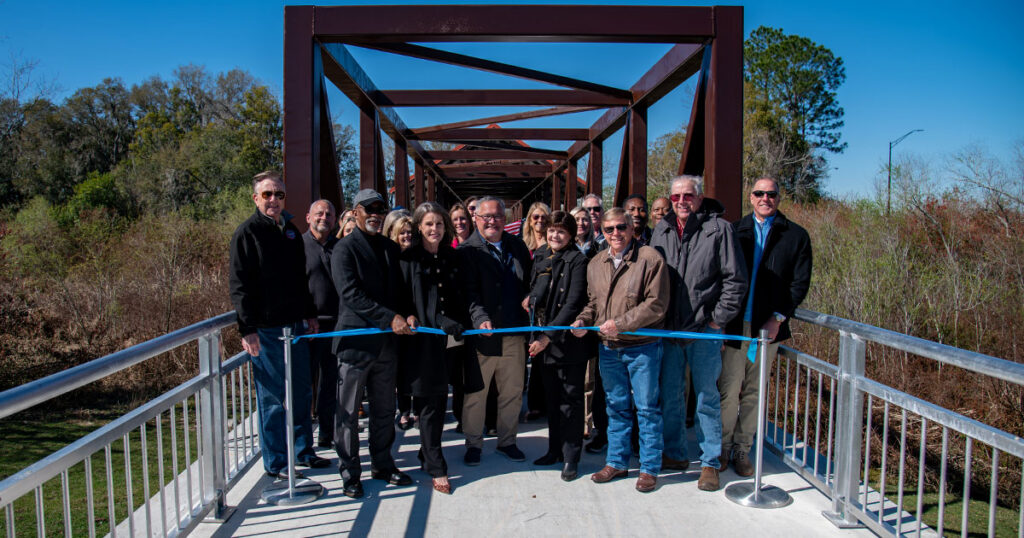 A group of people stands on a bridge for a ribbon-cutting ceremony. They hold a blue ribbon, smiling on a sunny day, with trees in the background.