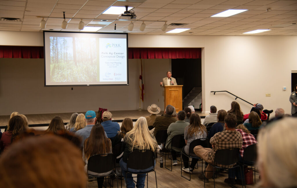 El concejal del condado de Polk, Rick Wilson, de pie en el escenario del evento, dirigiéndose a los asistentes sentados que acudieron a la reunión pública.