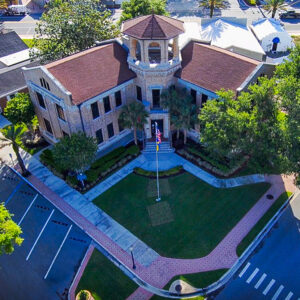 An aerial view of a historic brick civic building with a central octagonal tower, red‑tiled roof, tall windows and a landscaped lawn featuring a flagpole and palm trees.