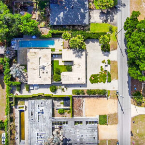 An aerial view of a modern residential property with a flat‑roofed home, central courtyard and long rectangular swimming pool, surrounded by hedges, trees and neighboring homes.