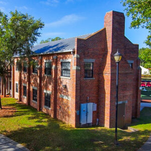 A two‑story brick armory building with a metal roof, tall rectangular windows and a prominent chimney‑like tower on one end. The structure sits beside a walkway with trees and open green space.