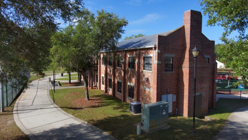 A two‑story brick armory building with a metal roof, tall rectangular windows and a prominent chimney‑like tower on one end. The structure sits beside a walkway with trees and open green space.
