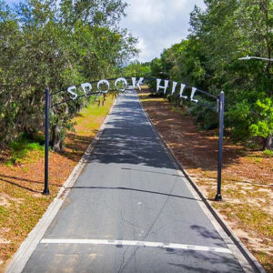 A paved road lined with trees leading uphill beneath a large metal archway that reads “Spook Hill.” The entrance is surrounded by natural vegetation and greenery.
