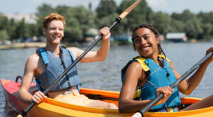 Two people wearing life jackets smile while kayaking together on a calm lake during a sunny day, paddling colorful kayaks with trees and shoreline visible in the background.