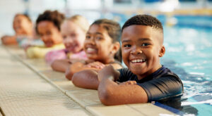 A diverse group of children smile while lined up along the edge of an indoor swimming pool during a swim lesson, with a young boy in the foreground leaning on the pool deck in a black swim shirt and other children visible behind him in the water.