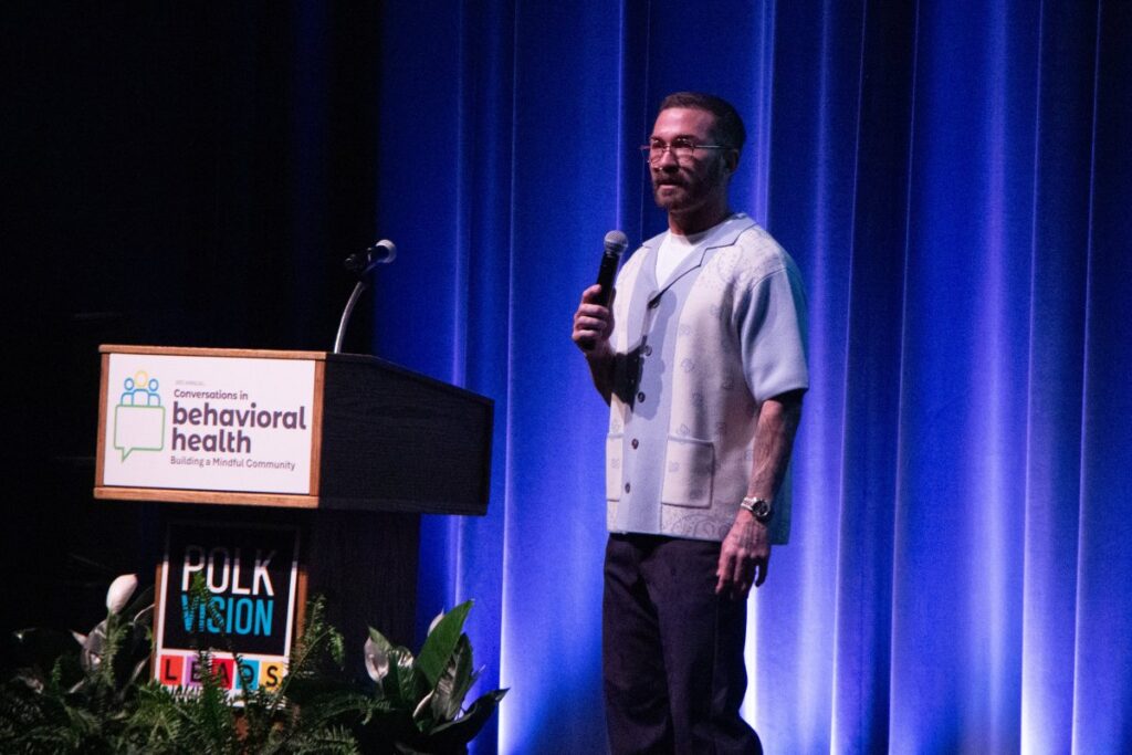 Tony Hoffman, a white man with short brown hair wearing glasses, stands on stage speaking into a microphone, a podium that says "conversations in behavioral health: building a mindful community" is behind him