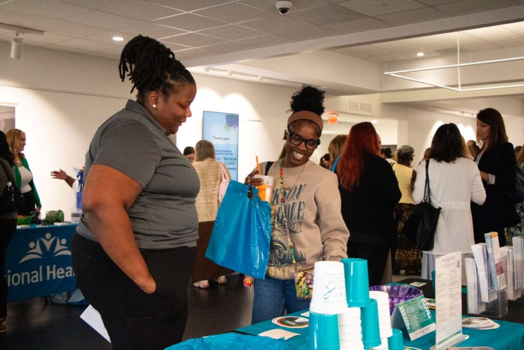 Two women interacting at an event booth with informational materials and stacked turquoise cups. Other people are visible chatting in the background.