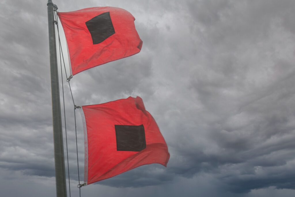 Two square red flags with small black squares in the center on a metal flag post flowing in the wind with gloomy dark clouds in the background