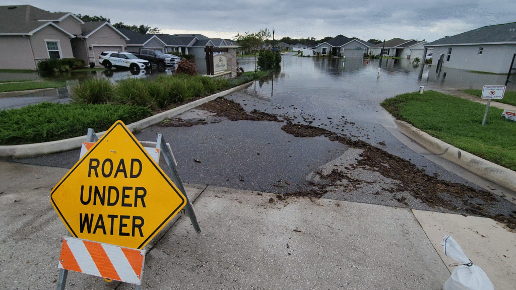 An orange, white and yellow metal roadway a-frame street sign that reads road under water placed on a sidewalk in front of a residential Polk County neighborhood entrance that is flooded with water in the background