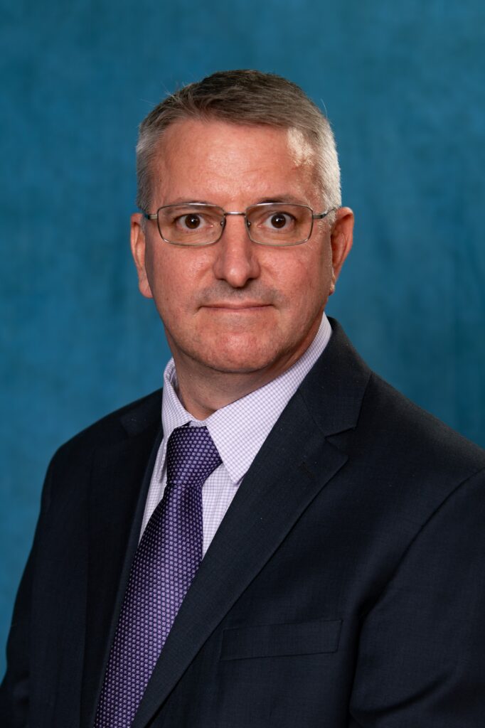 Headshot photo of next polk county real estate services division director scott lowery wearing a button up oxford collar shirt dark navy blazer purple pattern tie in front of a blue backdrop