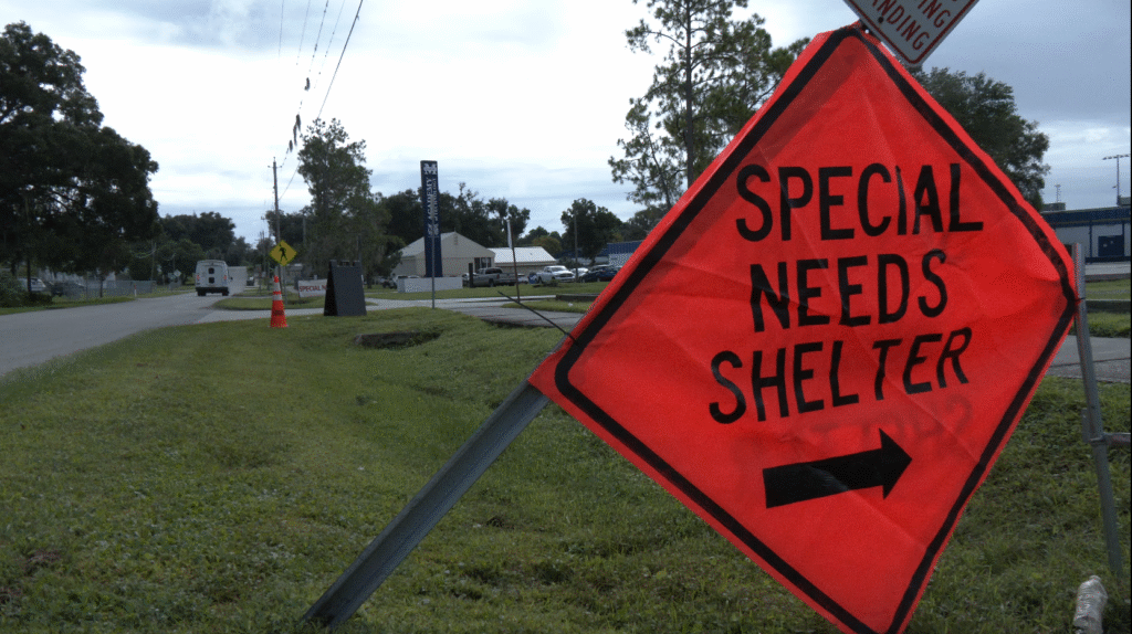 Photo image of a neon orange square roadway caution sign that reads special needs shelter with a right hand arrow in black writing on it strung from a metal post on the right side of a roadway with the entrance to McKeel Academy of Technology school in Lakland with white van traveling down roadway and vehicles parked in the school parking lot in the background