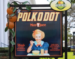 Decorative sign for the Polk County Citrus Label Tour featuring the vintage "Polkodot" citrus crate label. The artwork shows a young girl eating breakfast with a glass of orange juice, surrounded by text promoting Florida oranges, grapefruit, and tangerines. The display is framed with sculpted oranges and green leaves and stands in a landscaped garden outside a historic building.