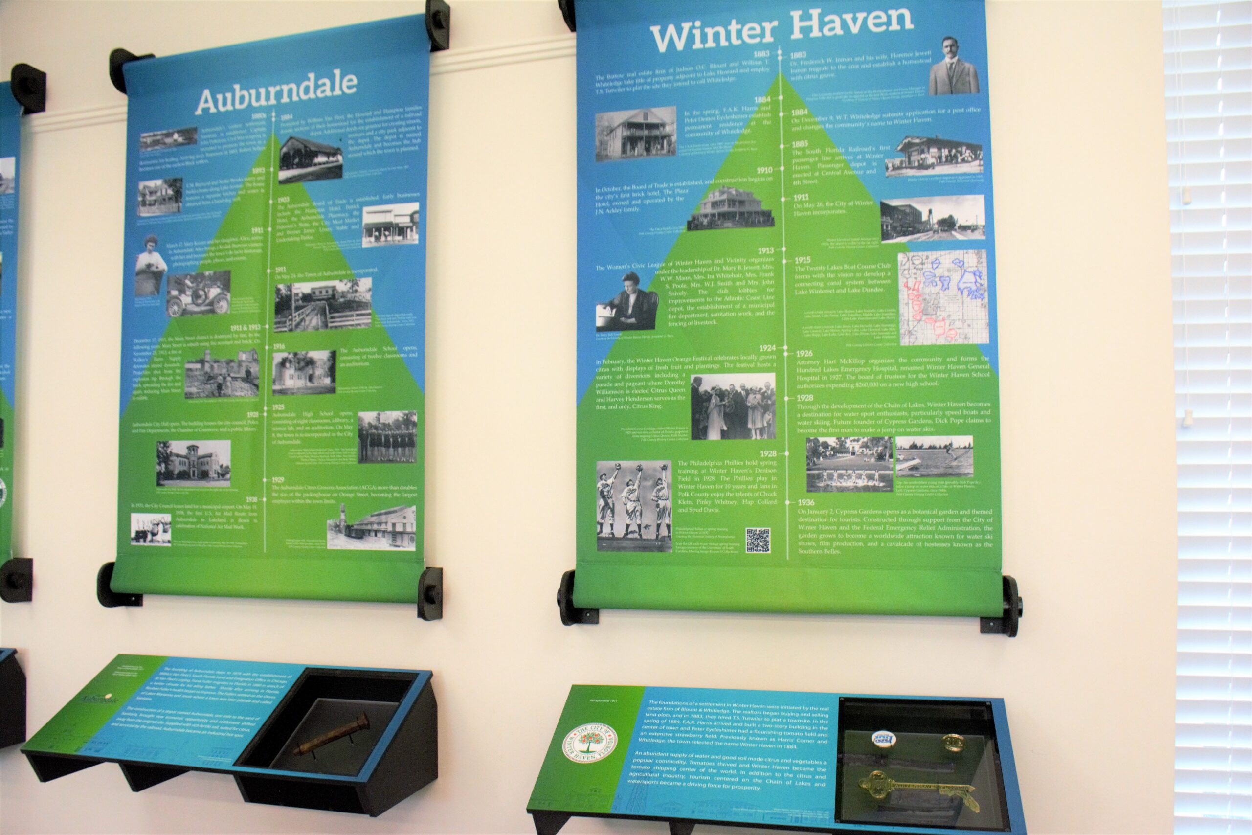 Timeline exhibit at the Polk History Center featuring illustrated panels highlighting the histories of Auburndale and Winter Haven. Historic photographs, milestone dates, and interpretive text trace the development of each city, with display cases below showcasing related artifacts and community information.