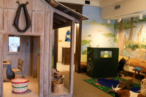 Children's interactive exhibit at the Polk History Center featuring a walk-through pioneer cabin with a hanging lantern, wooden furnishings, and hands-on play areas. A colorful mural of a Florida wetland with wildlife, including an alligator and birds, forms the backdrop, with activity stations and seating designed for young visitors.