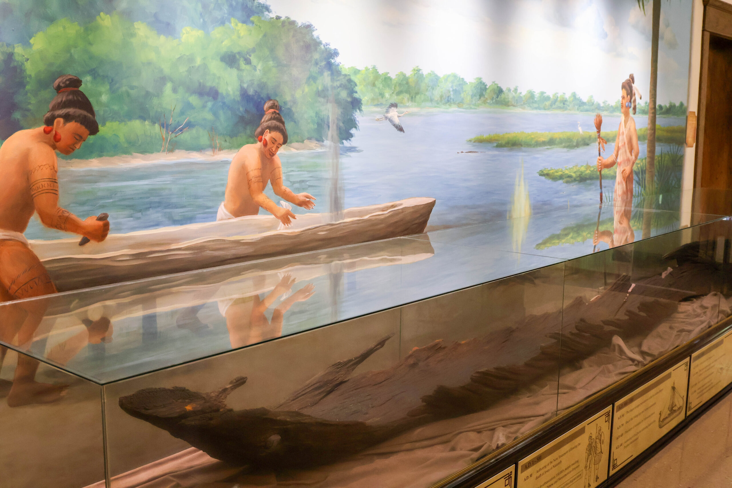 Exhibit at the Polk History Center featuring a mural of Florida's early Indigenous people carving a dugout canoe along a river. A preserved dugout canoe is displayed in a glass case in front of the mural, illustrating the craftsmanship and daily life of Florida's first inhabitants.
