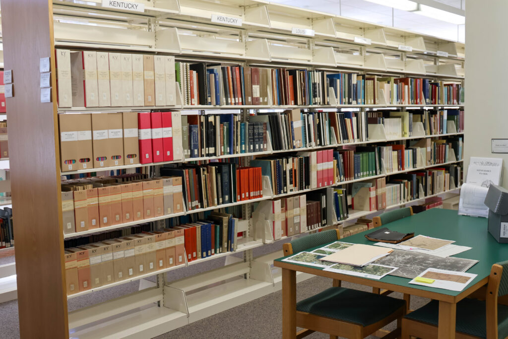 Rows of bookshelves filled with genealogy books, family histories, and archival reference materials in the Polk County History Center's Genealogical Library. A research table in the foreground holds maps, historic photographs, and documents for visitors to examine.