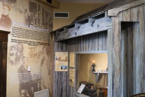 interior view of the Polk History Center's Pioneer Gallery featuring a recreated wooden pioneer cabin with a corrugated metal roof. Interpretive panels and artifacts, including an oil lamp, cookware, and household items, illustrate daily life of Polk County's early settlers. A large wall graphic titled 
