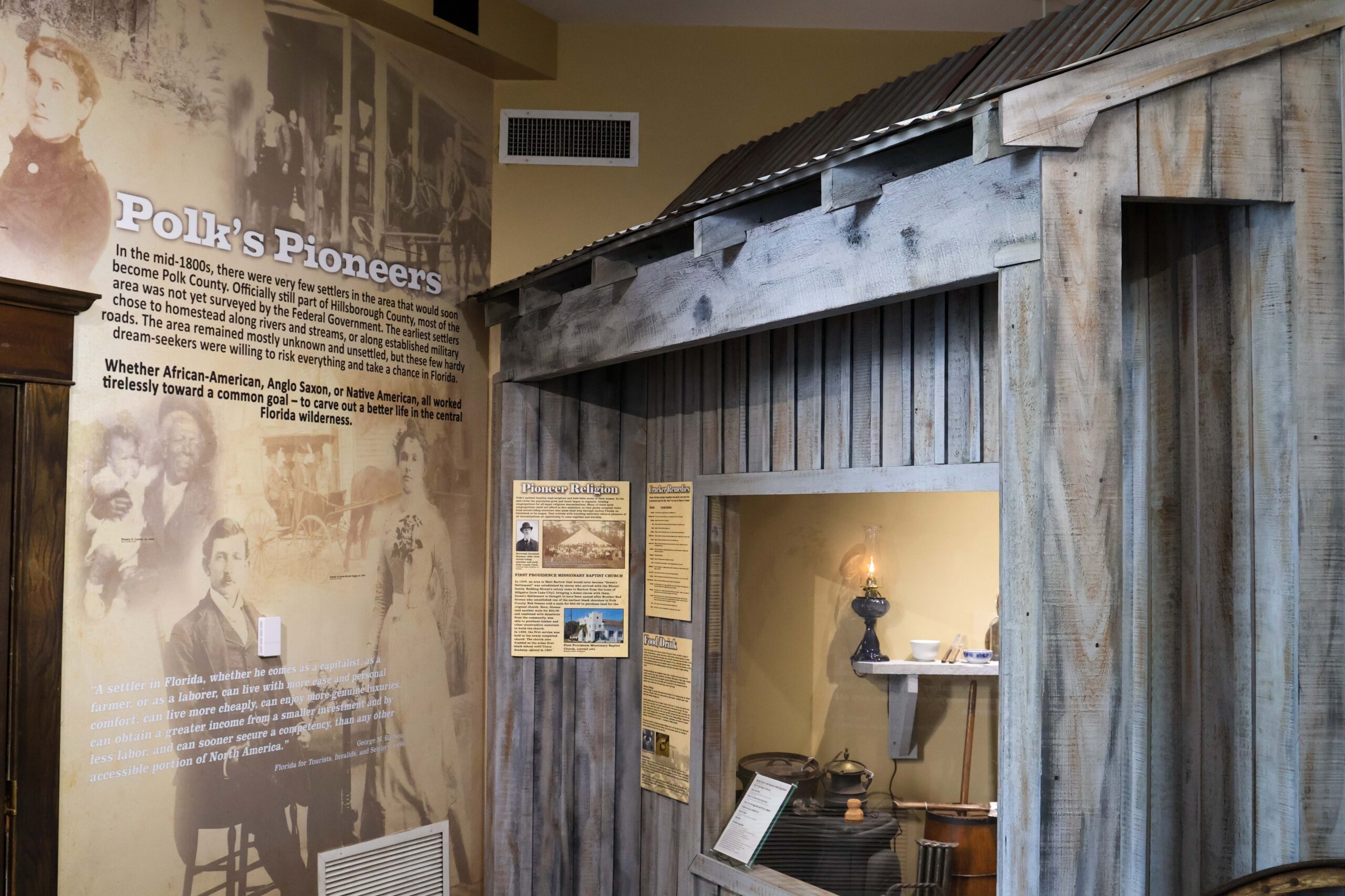 interior view of the Polk History Center's Pioneer Gallery featuring a recreated wooden pioneer cabin with a corrugated metal roof. Interpretive panels and artifacts, including an oil lamp, cookware, and household items, illustrate daily life of Polk County's early settlers. A large wall graphic titled 