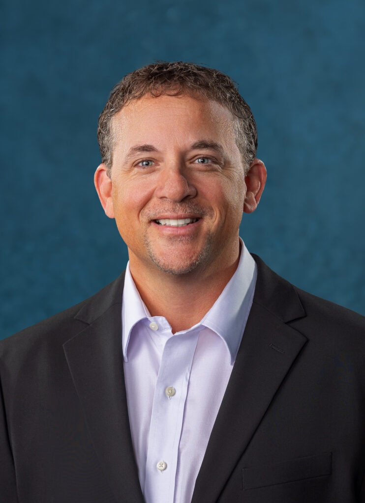 Ryan Neal Duncan smiles for a professional headshot, wearing a dark suit jacket and light blue shirt against a blue background.