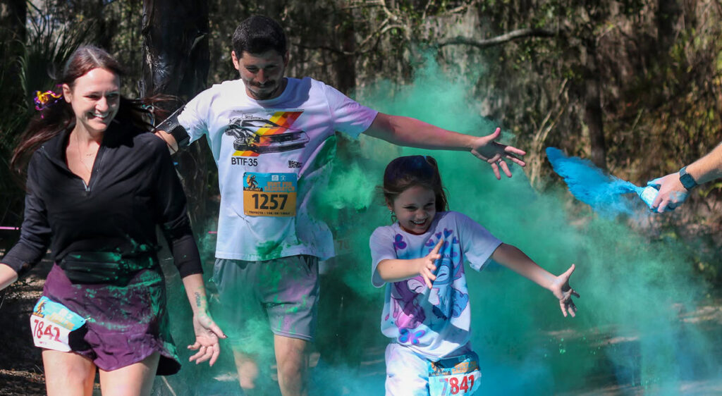 Tres participantes de una carrera de colores corren alegremente cubiertos de polvo verde, mientras uno de ellos lanza polvo azul al aire.