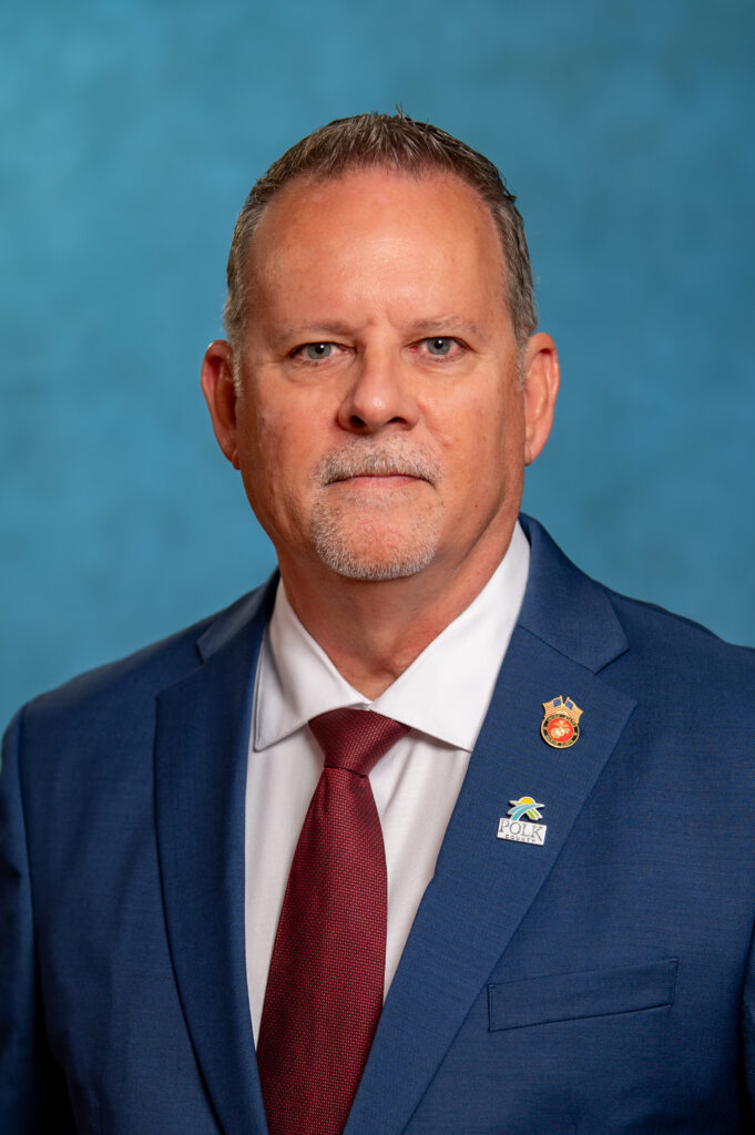 Official headshot of Jerry Rodriguez, the new Polk County Building Division Director. Rodriguez faces the camera against a solid blue background, wearing a navy blue suit jacket, white dress shirt and dark red tie. Two pins are displayed on his left lapel, including a Polk County logo pin. Rodriguez has short brown-and-gray hair and a neatly trimmed mustache and goatee.
