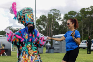A performer in a brightly colored traditional costume and feathered headdress holds hands with a woman in a blue shirt as they dance together outdoors.