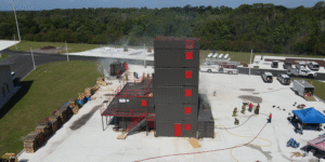 Aerial view of the Polk County Fire Rescue Training Center, showing firefighters practicing emergency response skills around a five-story training tower. Fire trucks, emergency vehicles, hoses and training equipment surround the tower, with wooded areas in the background.